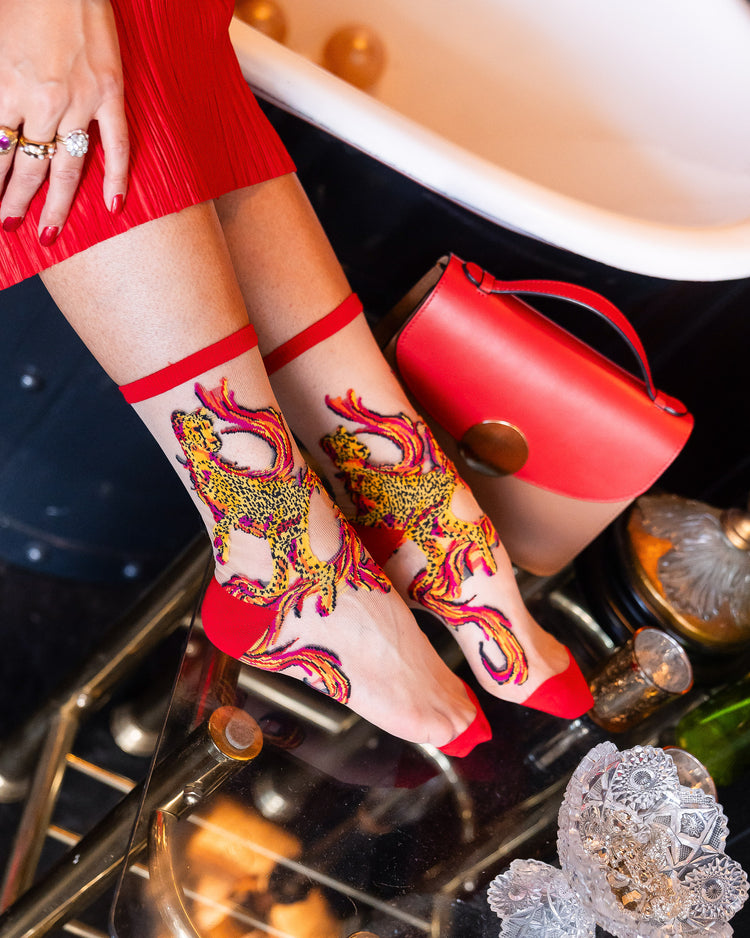 A woman wearing Sock Candy sheer socks with a red, orange, and yellow cheetah and flames motif with a red handbag in the background.