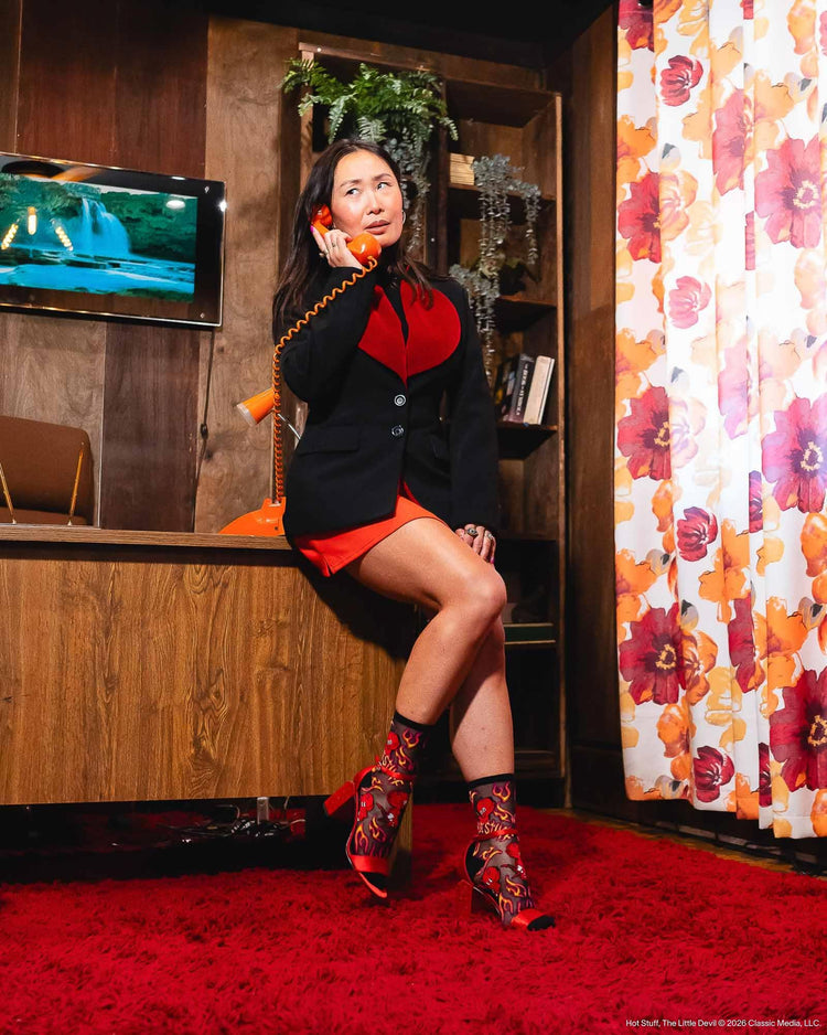 A woman posing in a retro lounge setting, wearing a black blazer with a red heart-shaped chest detail, a red mini skirt, and Sock Candy black sheer socks with a devil and flame design, paired with red strappy high-heel sandals . 