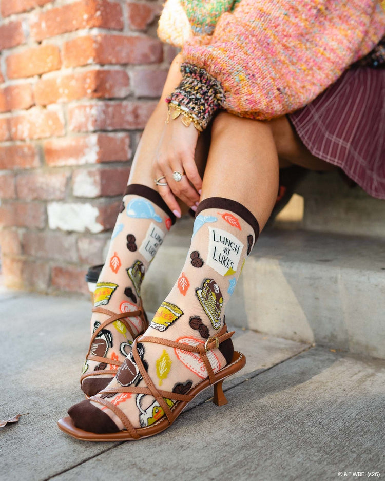 A close up Sock Candy Gilmore Girls Luke's Diner brown sheer crew socks decorated with coffee pots and autumn leaves, styled with open brown leather sandals.