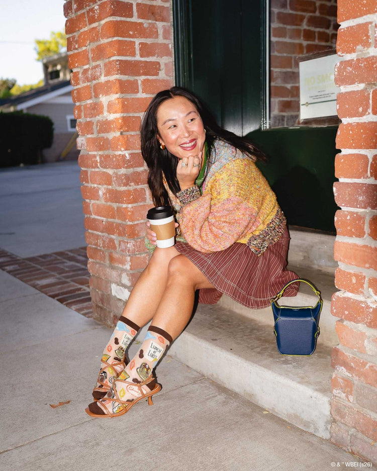 A woman wearing Sock Candy Gilmore Girls Luke's Diner brown sheer crew socks decorated with coffee pots and autumn leaves, styled with open brown leather sandals.