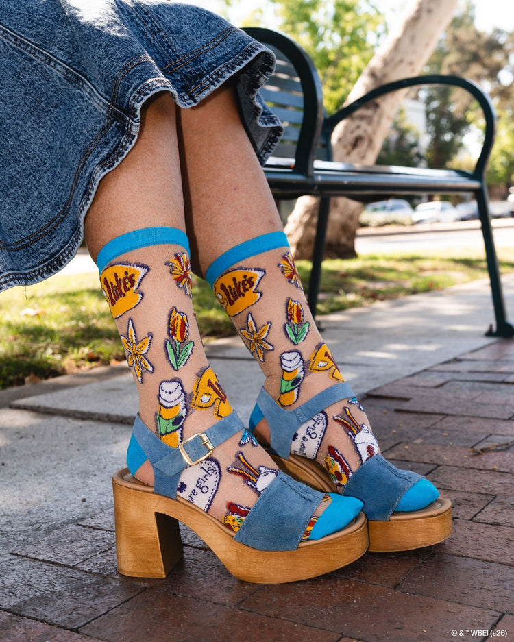 A woman wearing Sock Candy Gilmore Girls Stars Hollow sheer socks with detailed town gazebo and daisy prints, styled beautifully in high wooden platform sandals.
