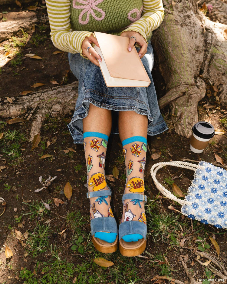 A woman wearing Sock Candy Gilmore Girls Stars Hollow sheer socks with detailed town gazebo and daisy prints, styled with high wooden platform sandals.