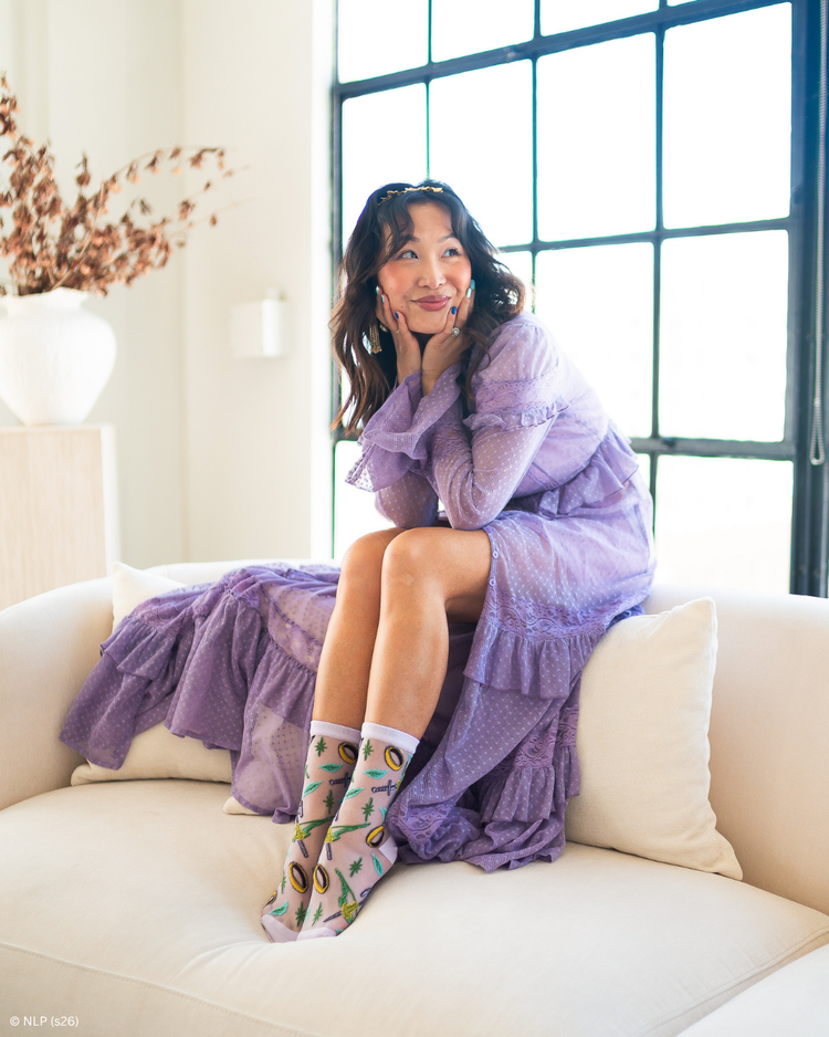 A woman sitting on a cream couch wearing a lilac sheer gown and a pair of sheer Sock Candy socks featuring a Lord of the Rings motif with a large window in the back.