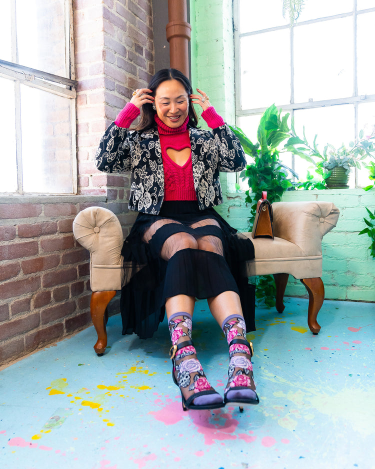 Woman sitting on a chair wearing sheer Sock Candy socks with a floral and skulls motif and black high-heeled sandals in a room with large windows and plants.