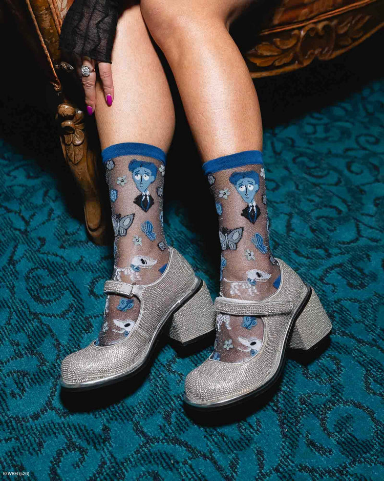 A woman seated in an ornate chair wearing sheer Sock Candy patterned socks with a design motif from Tim Burton's The Corpse Bride, paired with glittery silver Mary Jane-style heels, on a teal patterned carpet.