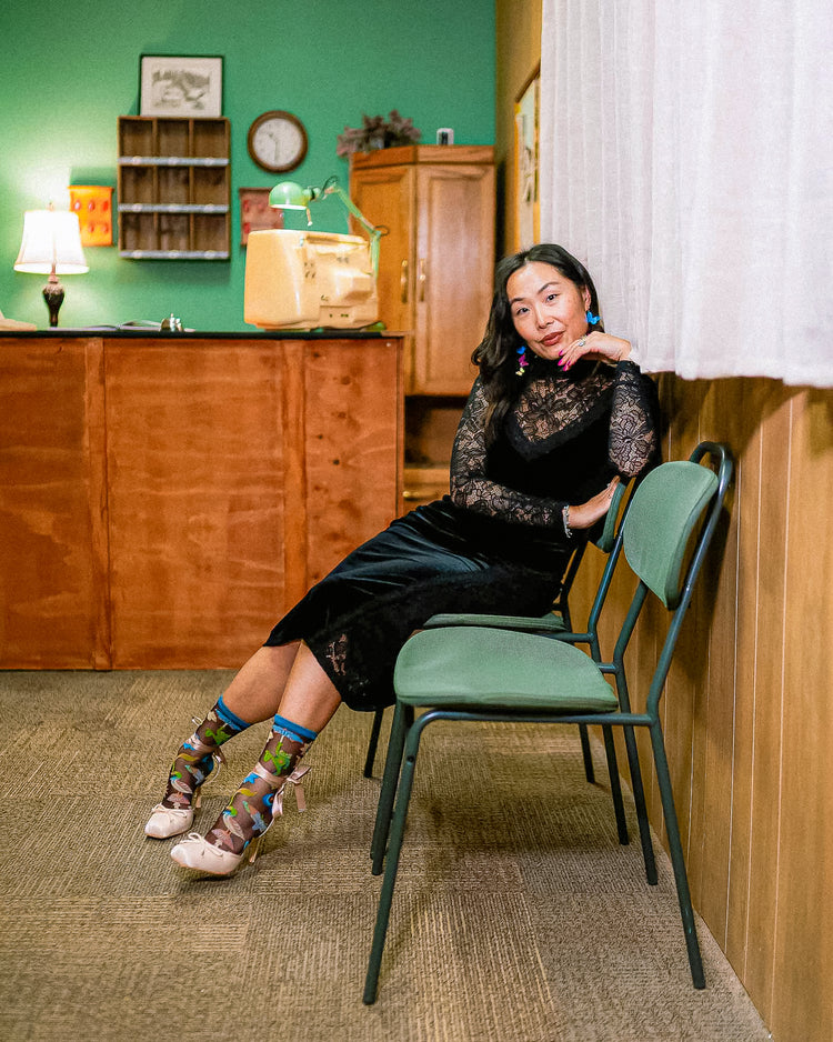 A woman in a black lace and velvet long-sleeve dress and sheer Sock Candy socks sits on a green chair in a vintage-style room with a wood-paneled wall and green wall in the background.