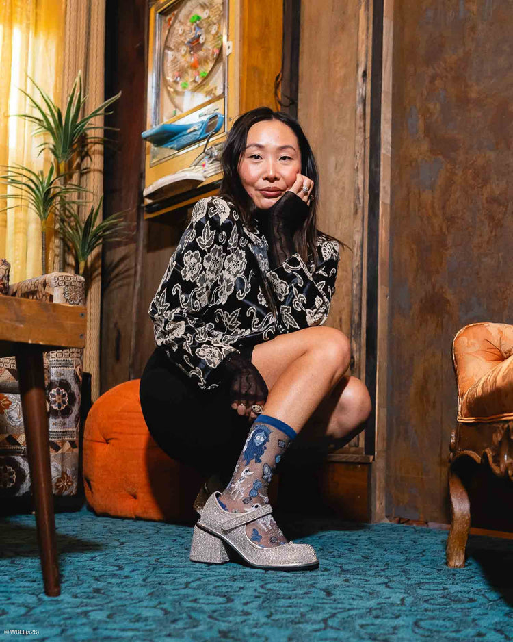 A woman crouches in a vintage-style room wearing a black and white paisley patterned blouse, black skirt, sheer Sock Candy socks, and sparkly silver Mary Jane shoes.