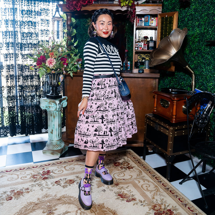 A woman stands in a vintage-themed room wearing a black and white striped long-sleeve top, a pink and black graphic print skirt,  purple Doc Martens shoes, and sheer Sock Candy black cat socks.