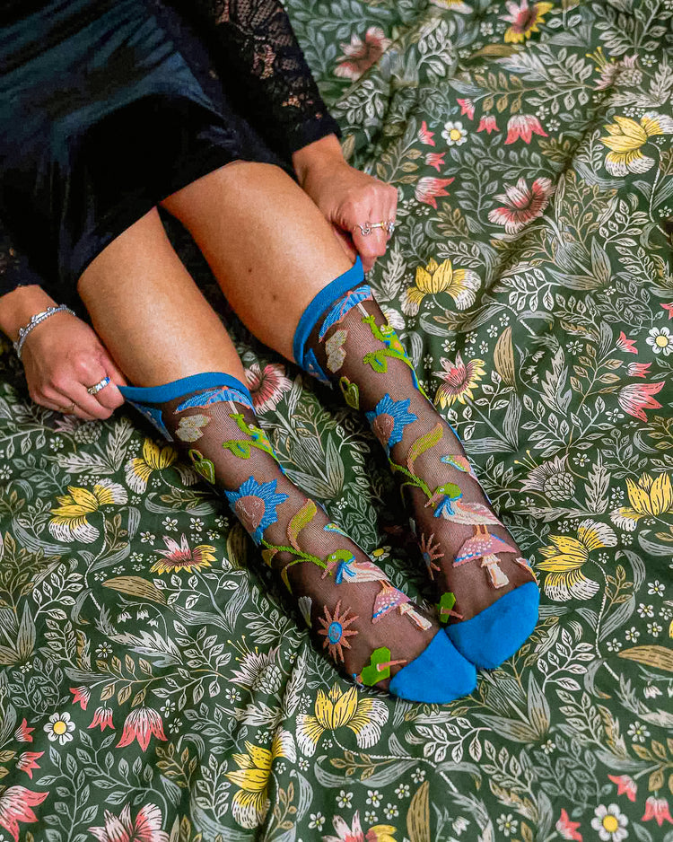 A woman wearing a pair of sheer Sock Candy socks with a colorful bird and floral print in a whimsical dark fairytale motif on an ornate green comforter.