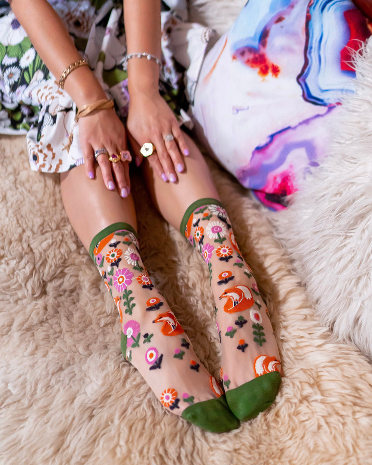 A woman sitting on a beige faux-fur rug wearing sheer Sock Candy socks with a colorful woodland print of foxes, flowers, and green cuffs and toes.