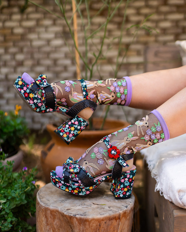 Colorful floral high-heeled shoes paired with sheer Sock Candy rabbit socks worn by a woman sitting on a wooden stump with a garden background.