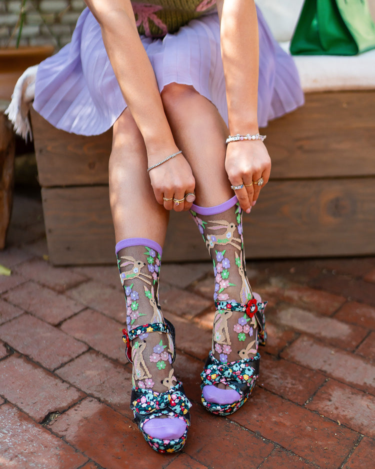A woman wearing floral embroidered shoes with black sheer Sock Candy rabbit socks on a brick pavement.