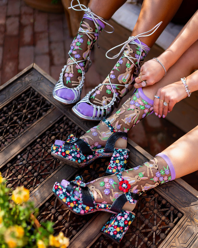 Two women wearing black sheer Sock Candy socks with a rabbit and floral motif, one paired with silver strappy high-heeled sandals and one pair with colorful floral heels.
