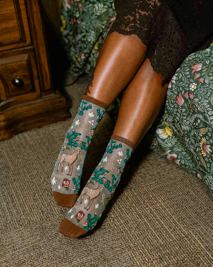 A woman seated on a patterned sofa wearing sheer Sock Candy socks with a deer and floral print on a shimmery background, with brown heels and toes.