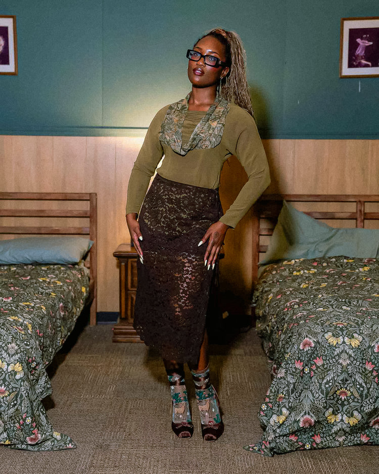 A woman wearing an olive green top and brown lace outfit paired with sheer Sock Candy deer motif socks and brown ballet heels stands in a bedroom.