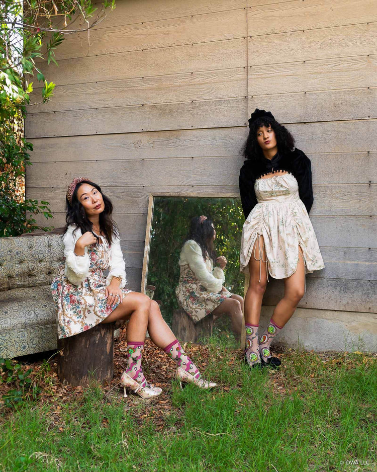 Two women posing outdoors against a wooden wall, wearing best friend style floral mini dresses in true Galentine's style, sheer Shrek fashion socks, and heels, with one seated on a rustic tree stump and one standing beside a mirror.