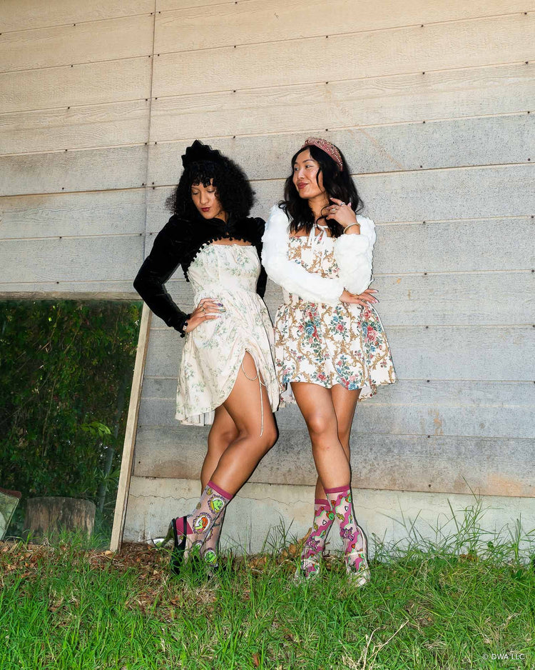 Two women posing outdoors against a weathered wood wall, wearing best friend style matching outfits with floral minidresses, sheer Shrek fashion socks and heels, standing on grass.