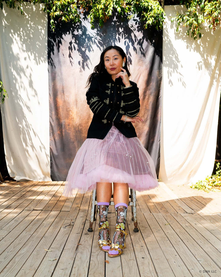A woman sitting on a metal stool on top of a wooden stage, with a dark backdrop and tree branches in the background. She is styled in an edgy feminine aesthetic and wearing Shrek black sheer fashion socks with metallic gold heeled sandals and a pink tulle petticoat skirt.