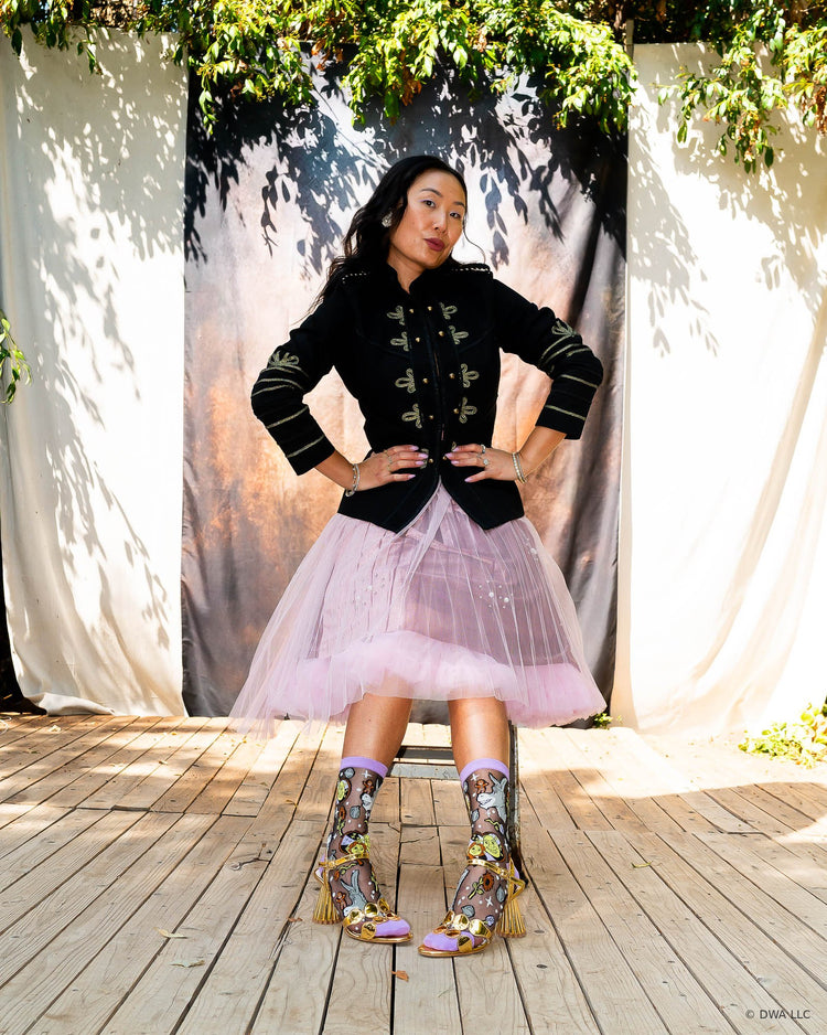 A model sitting on a metal stool on a wooden stage in front of a dark backdrop, wearing black sheer Shrek fashion socks with gold metallic heels and an edgy feminine outfit consisting of a black military jacket and pink tutu skirt.