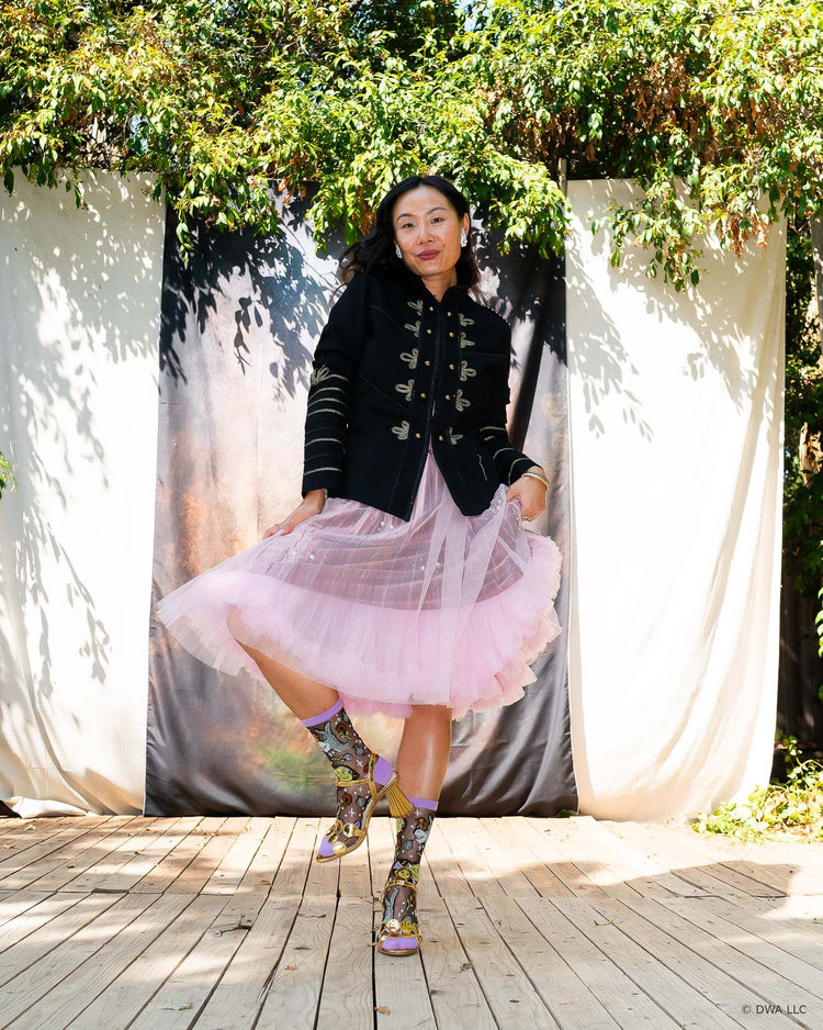 A woman standing on a wooden stage in front of a dark backdrop and surrounded by nature, posing in a pair of Shrek black sheer fashion socks with metallic gold heeled sandals and styled in an edgy feminine aesthetic with a pink tulle skirt and black military style jacket. 