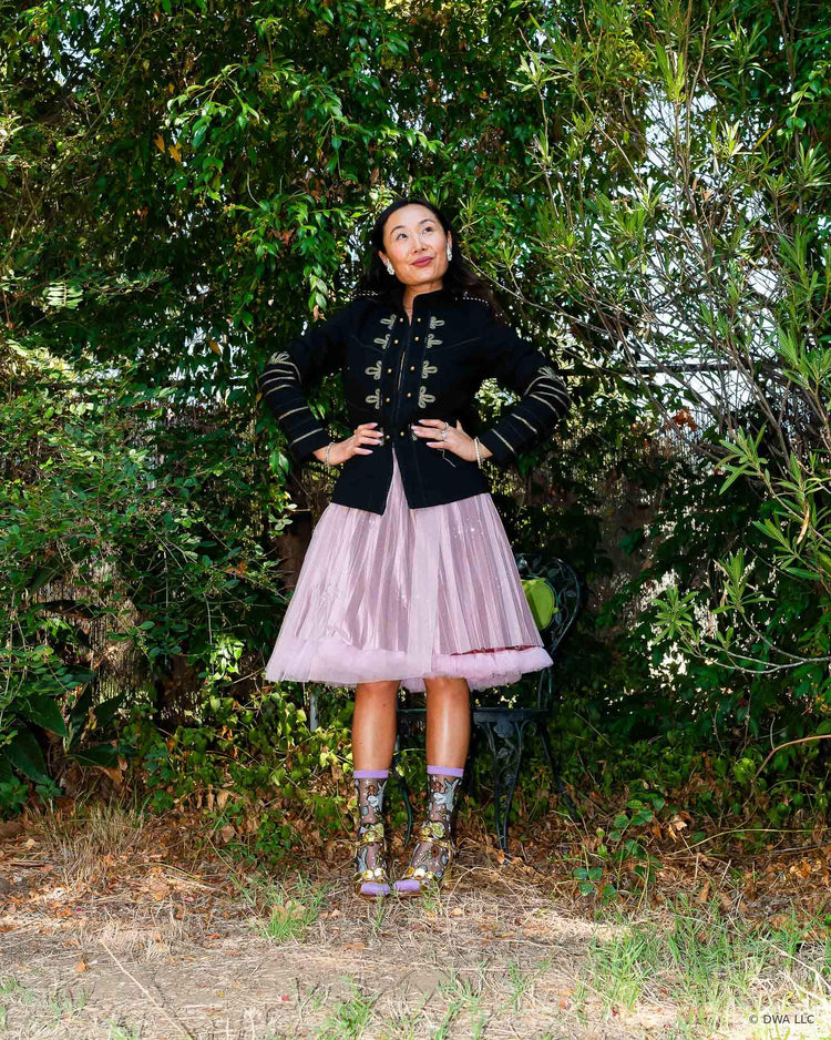 A woman standing in nature with her hands on her hips, styled in an edgy feminine aesthetic with a black military style jacket and pink tulle petticoat skirt. She is wearing Shrek black sheer fashion socks and metallic gold heeled sandal.