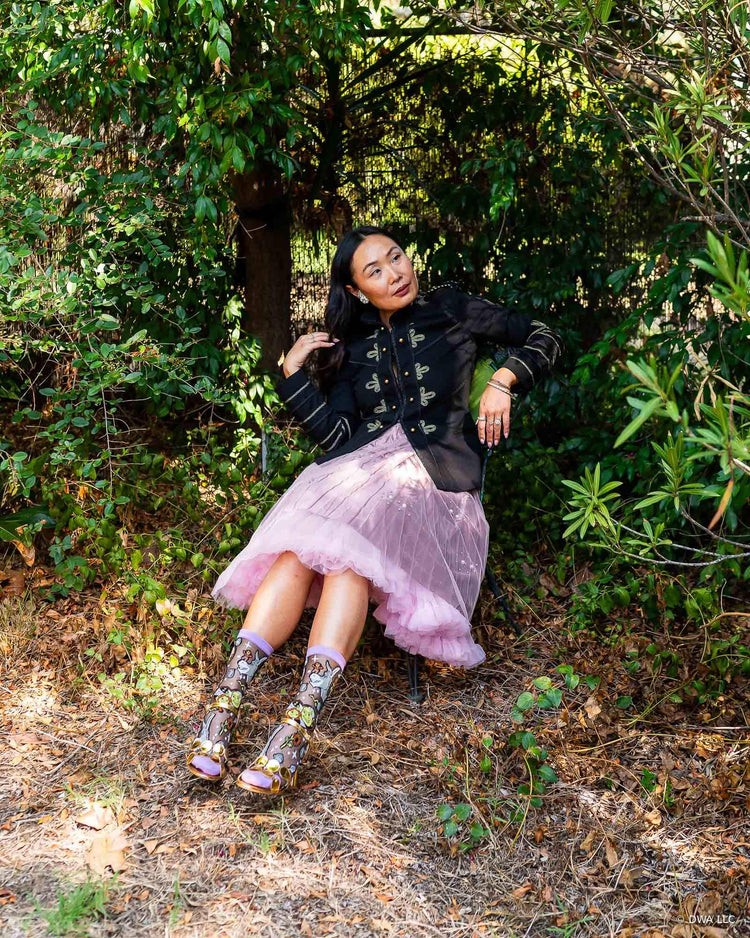 A woman sitting on a metal chair surrounded by nature, wearing a black military style jacket and pink tulle skirt for edgy feminine styling. She is wearing Shrek black sheer fashion socks designed by Sock Candy and metallic gold heeled sandals.