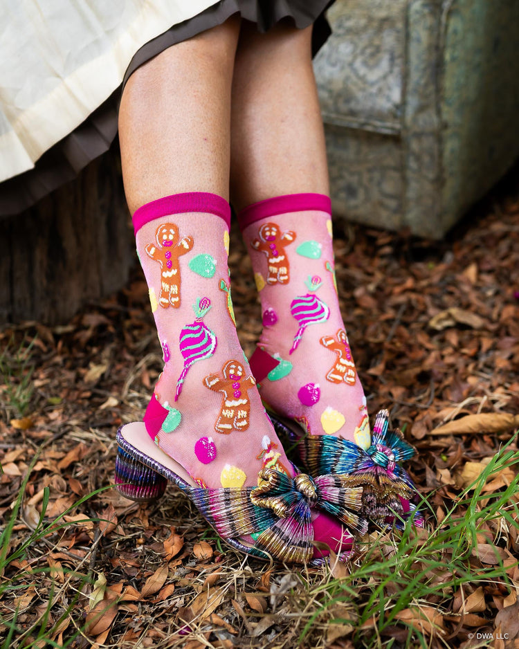 A pair of Shrek sheer pink Christmas fashion socks with a colorful candy and gingerbread design, worn with metallic bow heels outdoors on a leaf-covered ground.