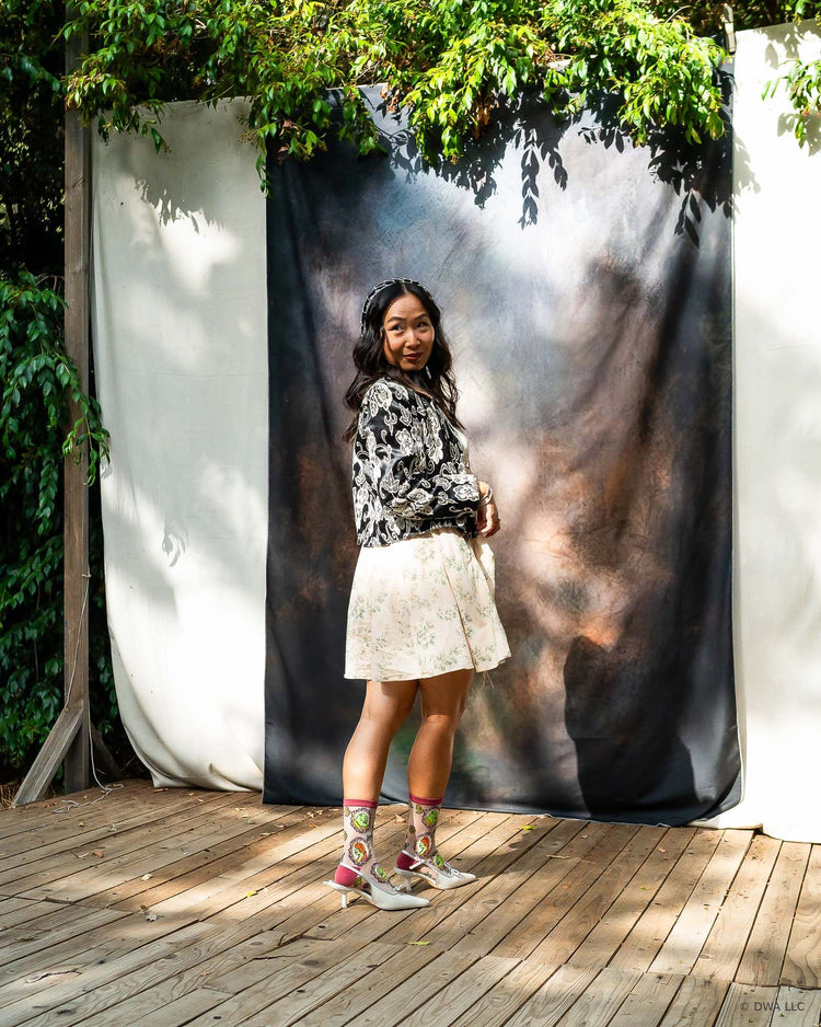 A model stands on a wooden stage in front of a dark backdrop, wearing a black and white patterned long-sleeve top, a cream floral dress, and sheer Shrek socks paired with slingback heels in a coquette aesthetic.