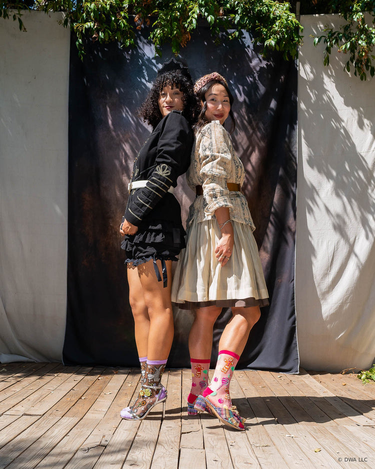 Two women posing together outdoors on a wooden stage in front of a draped black backdrop with leafy branches overhead. The woman on the left wears a high fashion black military jacket and bloomers, and the woman on the right wears a cream-colored patterned jacket and flowy skirt. They are both wearing sheer Shrek holiday socks with heels.