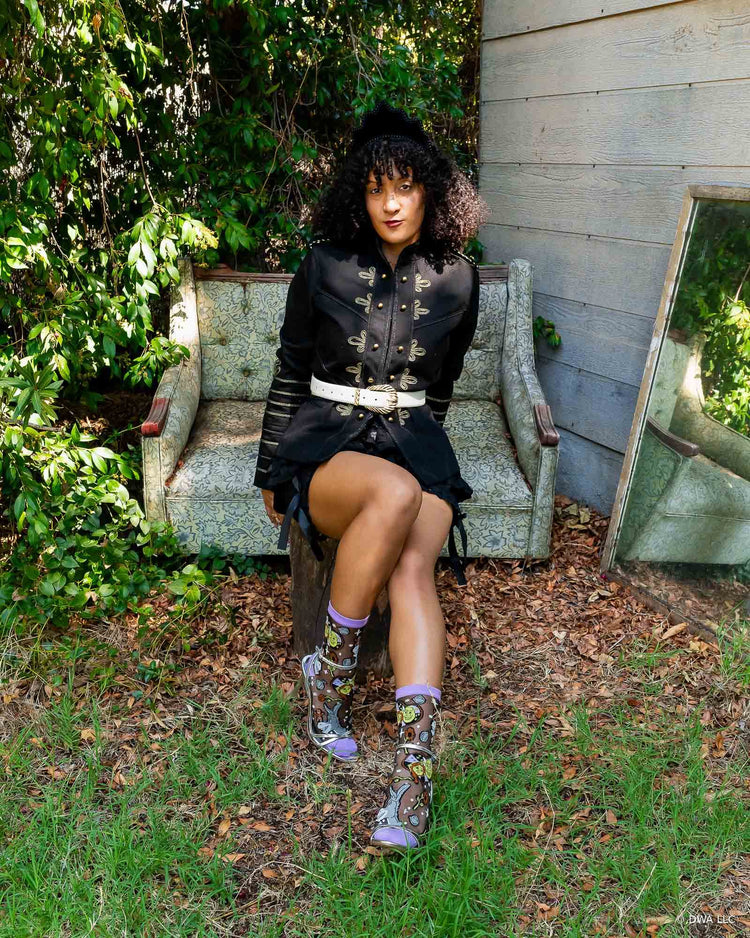 A model sitting on a vintage couch outdoors, wearing a high fashion black military jacket, black bloomers, and sheer Shrek socks designed by Sock Candy with silver metallic heels. 