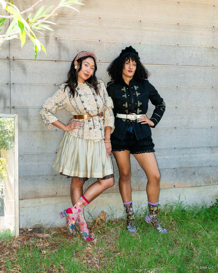 Two women posing outdoors against a rustic gray wooden wall, wearing bohemian-style outfits. The woman on the left wears a beige floral long-sleeve top and pleated skirt and the woman on the right wears a high fashion black military jacket with bloomers. Both are wearing sheer Shrek holiday socks with heels.