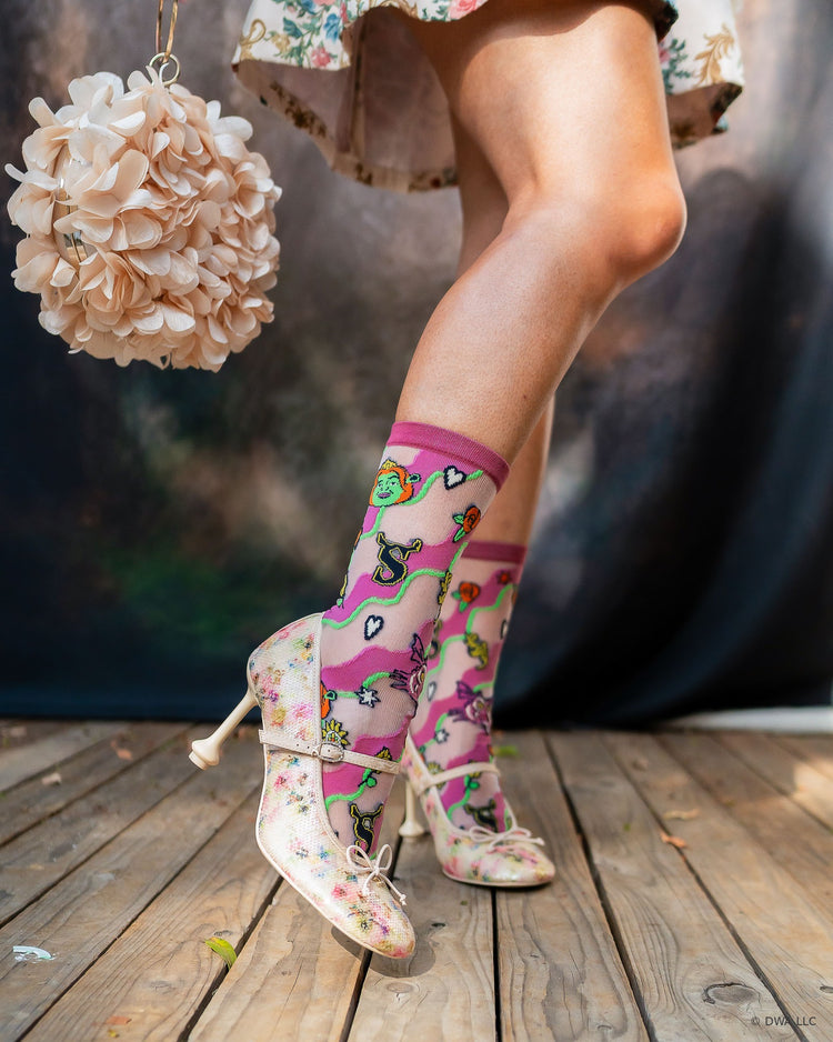 A woman's legs wearing sheer Shrek high fashion socks designed by Sock Candy, paired with floral Mary Jane heels, standing on a wooden stage with a floral hanging bag nearby.