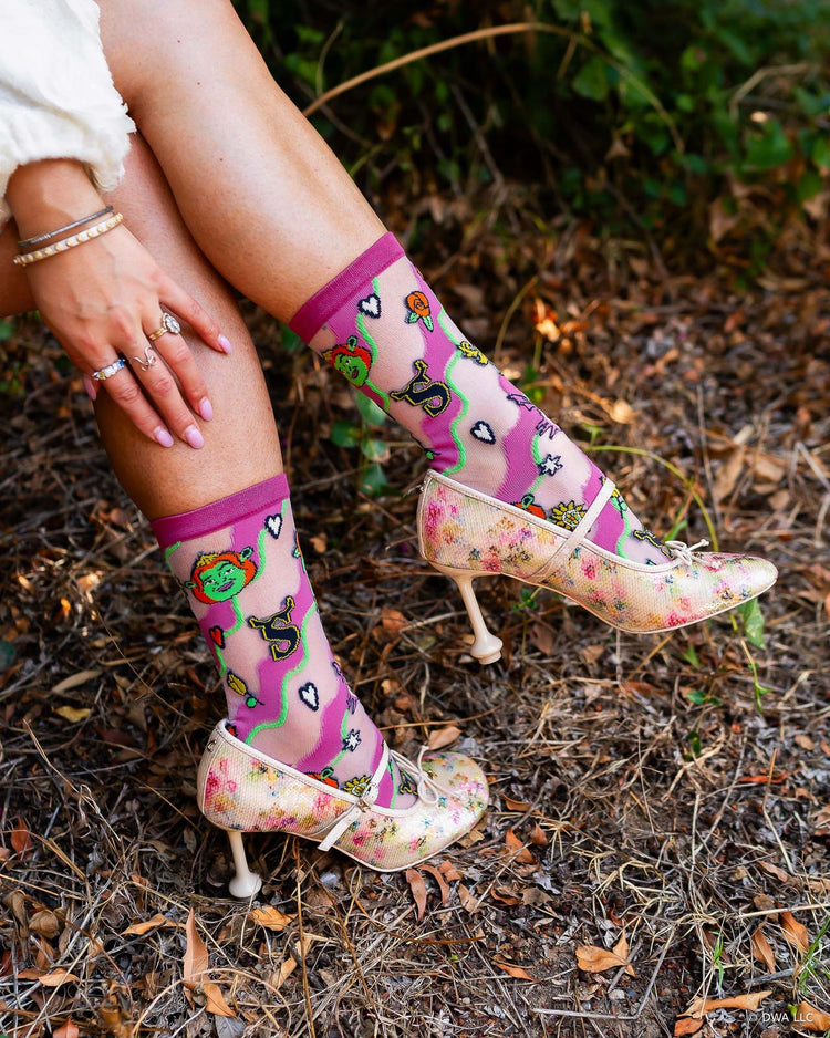 A woman sitting outdoors wearing high fashion sheer Shrek socks from Sock Candy with a whimsical print paired with floral Mary Jane heels.