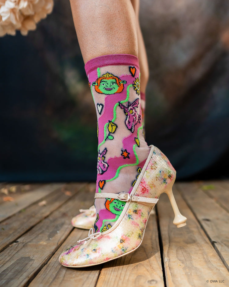 A woman wearing sheer high fashion Shrek socks featuring Princess Fiona and Dragon, paired with floral high-heeled Mary Jane shoes, standing on a wooden stage.
