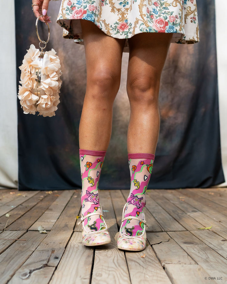 A close-up of a model's legs and feet standing on a wooden stage with a dark backdrop in the background, wearing high fashion sheer Shrek socks with floral Mary Jane heels paired with a floral minidress.