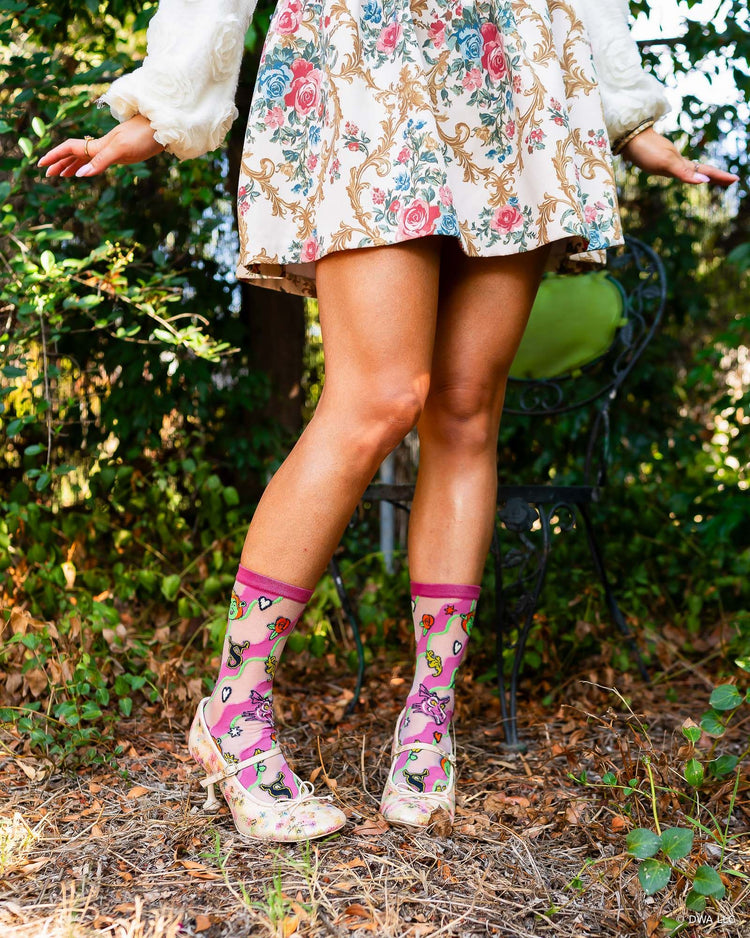 A woman  outdoors wearing a cream floral dress and sheer high fashion Shrek socks paired with floral Mary Jane heels. The background shows green foliage and a black garden chair.