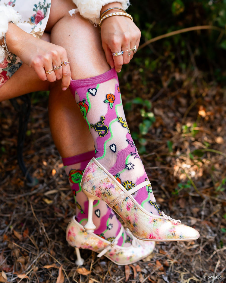 A woman outdoors wearing floral Mary Jane heels and high fashion sheer Shrek socks designed by Sock Candy, seated on a bench with leaves and greenery in the background.