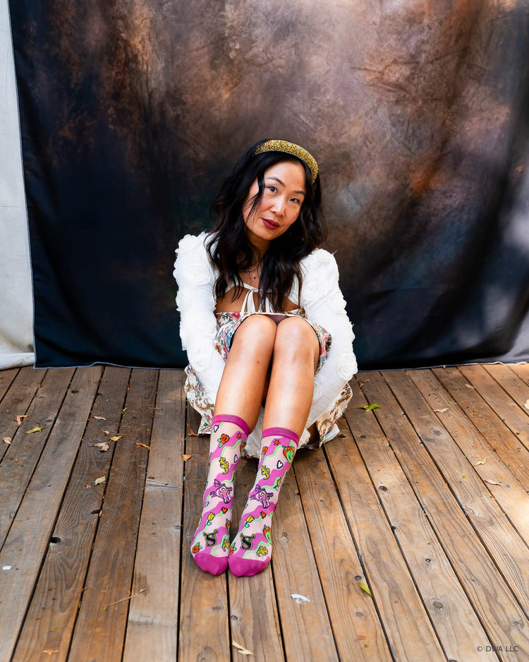 A woman sits on a wooden stage in front of a dark backdrop, wearing a white fuzzy cardigan, a floral coquette style minidress, and sheer pink Shrek socks featuring a Princess Fiona and Dragon design motif.
