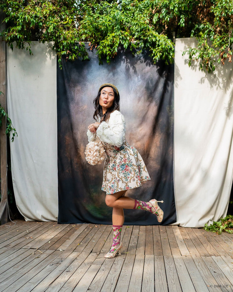A woman posing outdoors in front of a printed backdrop, wearing sheer pink Shrek socks with a floral coquette style dress, paired with floral Mary Jane high heels.