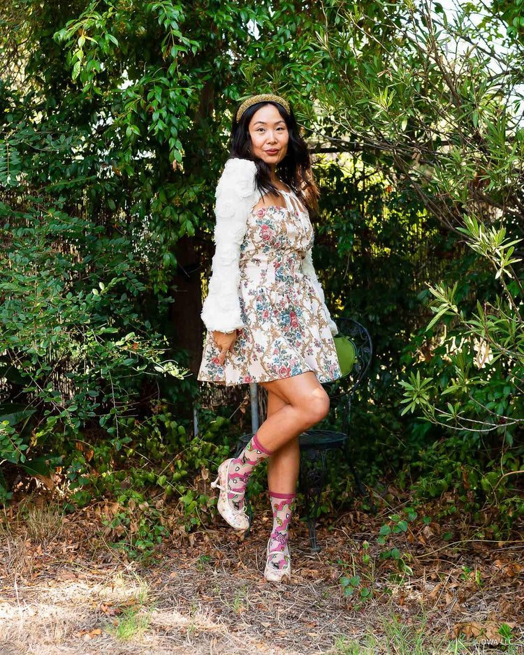 A woman posing outdoors in a garden setting, wearing a short white jacket over a floral mini dress in coquette styling, paired with sheer pink Shrek socks and floral Mary Jane heels.
