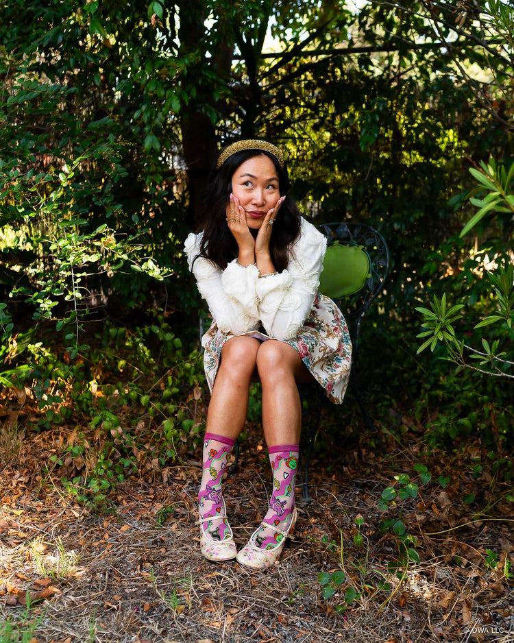 A woman sitting on a metal chair outdoors in a wooded area, styled in a coquette floral minidress, sheer pink Shrek socks, and a pair of Mary Jane high heels.