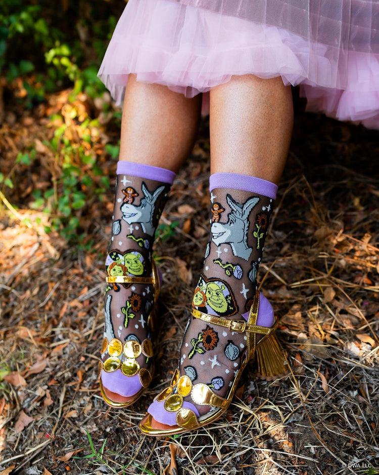 A close up of women's legs in an outdoor setting wearing sheer black Shrek socks paired with gold metallic heeled sandals and a pink tutu.