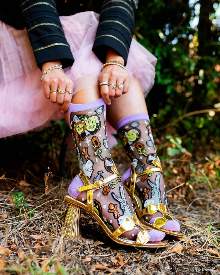 A woman seated outdoors wearing black sheer Shrek fashion socks with a colorful cartoon print, paired with metallic gold high-heeled sandals.