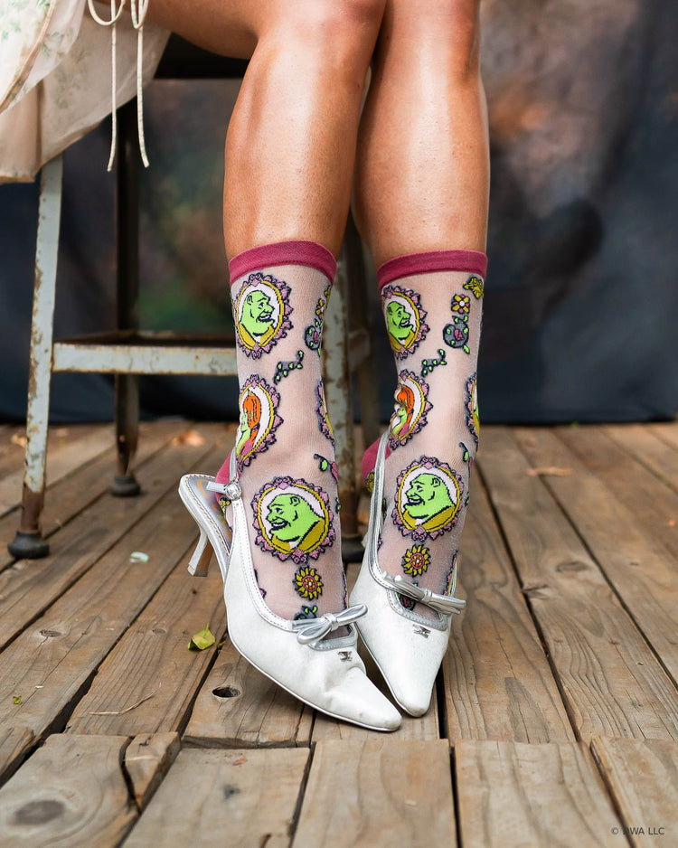 A close-up of a women's legs and feet sitting on a metal stool on a wooden stage. She is wearing sheer Shrek fashion socks paired with slingback heels.