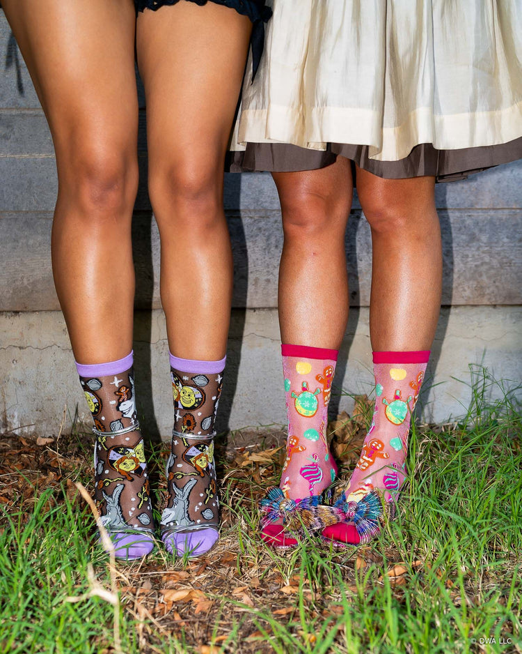 Two women standing outdoors on grass in front of a wooden wall wearing sheer Shrek holiday socks with metallic heels in a festive style.