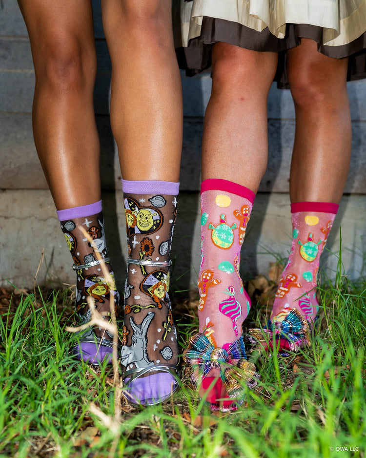 A close-up of two women's legs standing on grass wearing sheer Shrek holiday socks designed by Sock Candy. The model on the left is wearing socks with metallic silver heels and the model on the right is wearing socks with metallic bow heels.