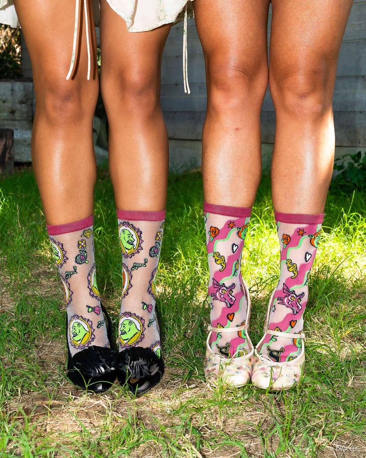 A close up of two women's legs on grass outdoors, wearing sheer Shrek fashion socks designed by Sock Candy. The socks are worn with a pair of pointy black heels and a pair of floral Mary Jane heels.