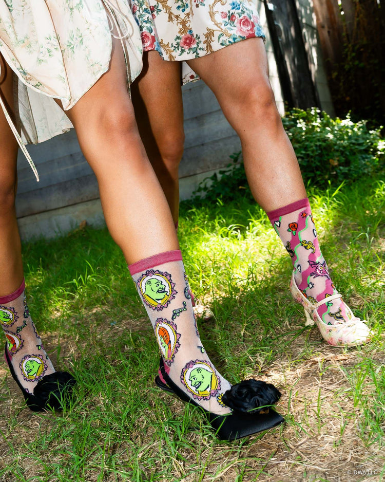A close-up lifestyle shot of two women walking on grass, wearing sheer Shrek fashion socks designed by Sock Candy paired with heels and floral dresses.