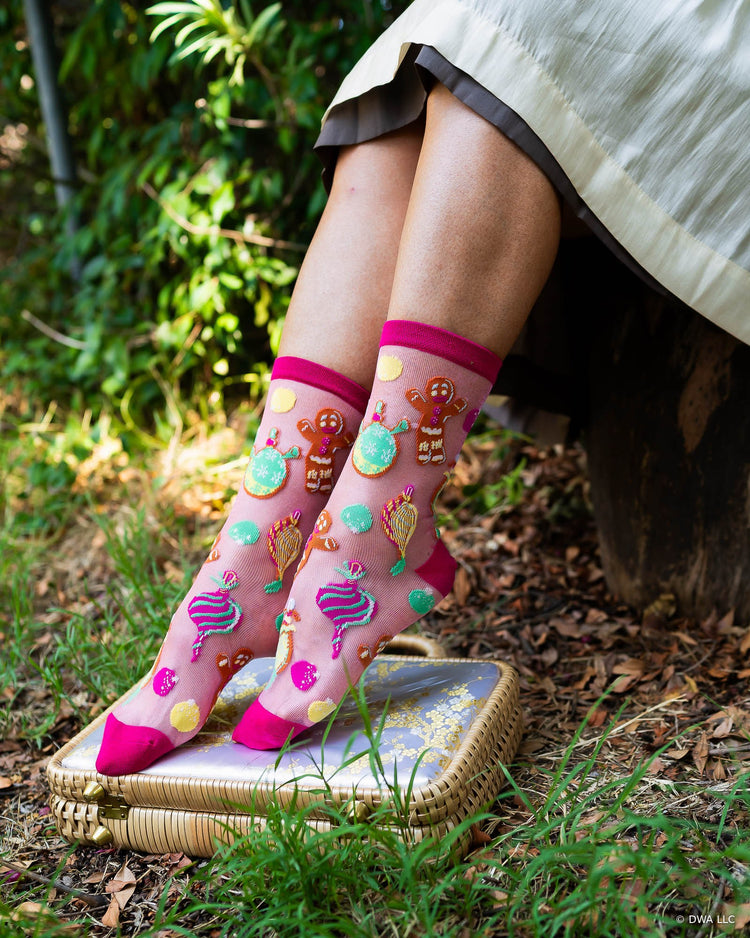 A woman sitting outdoors on a tree stump wearing Shrek sheer pink holiday socks with colorful gingerbread-themed illustrations. The background shows green grass and foliage.