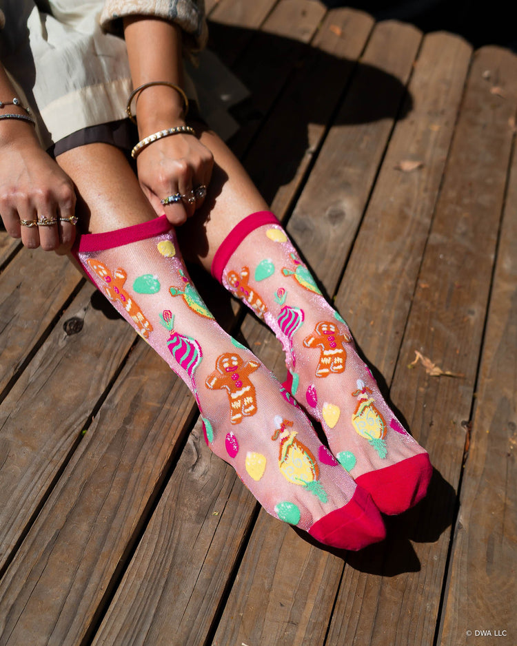 A woman sitting on a wooden stage wearing sheer Shrek pink socks with a colorful candy and gingerbread pattern designed by Sock Candy.
